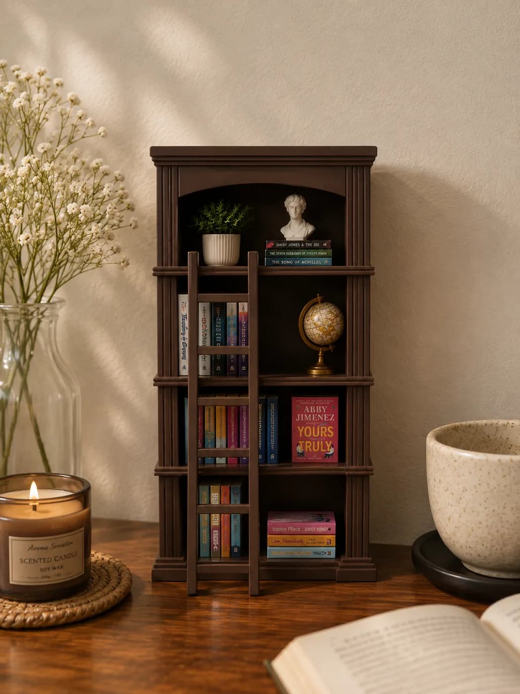 A choco brown miniature bookshelf filled with tiny books, a plant and a brass globe, lit by window light beside a candle and an open book