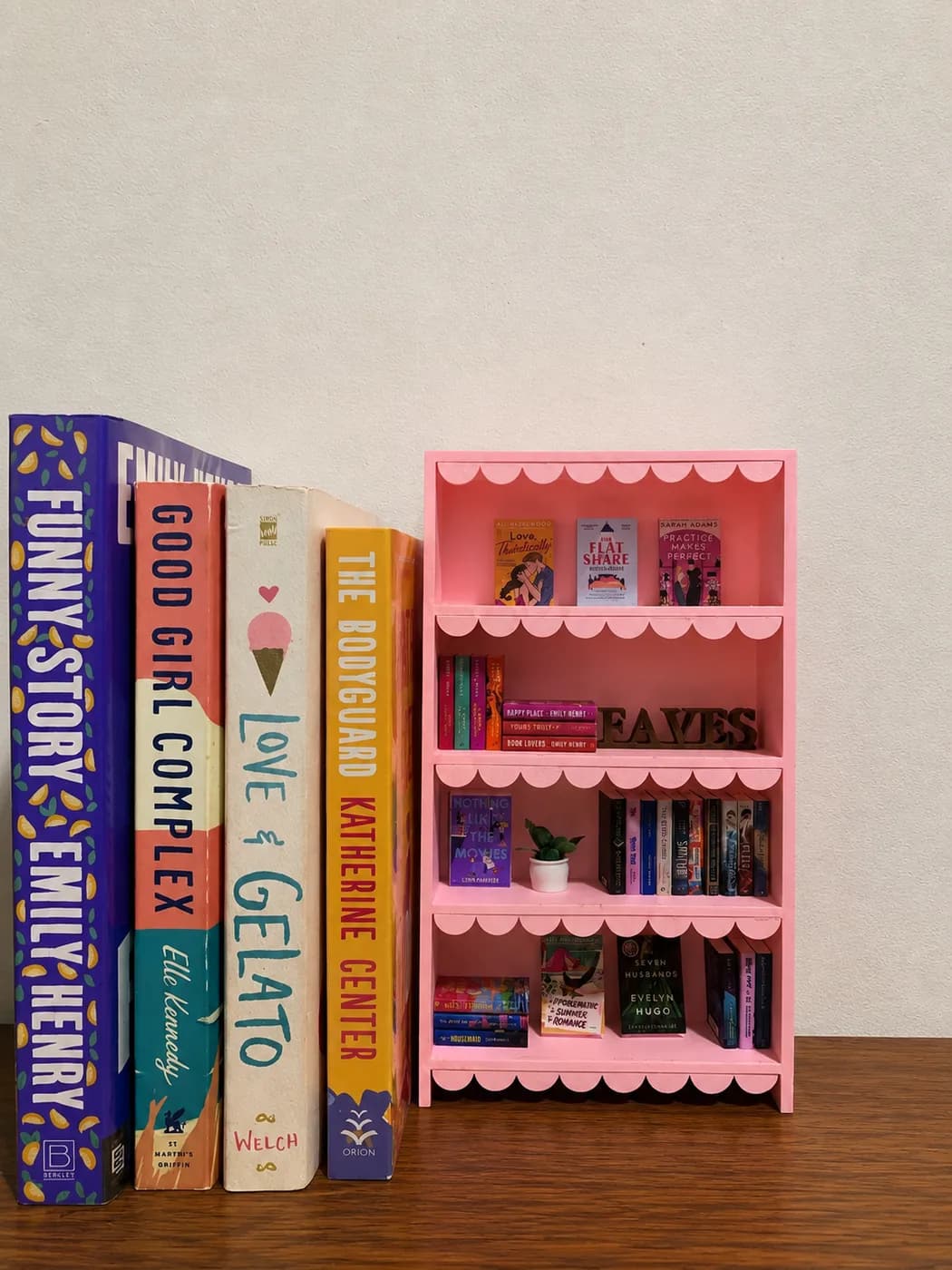 A Blush Pink Scalloped bookshelf filled with tiny hardcovers and wooden FAVES letters, standing beside full-size paperbacks